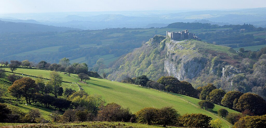Carreg Cennen Castle: A View of a historic stone castle ruins on top of a rocky hill overlooking rolling green fields and wooded areas under a clear sky