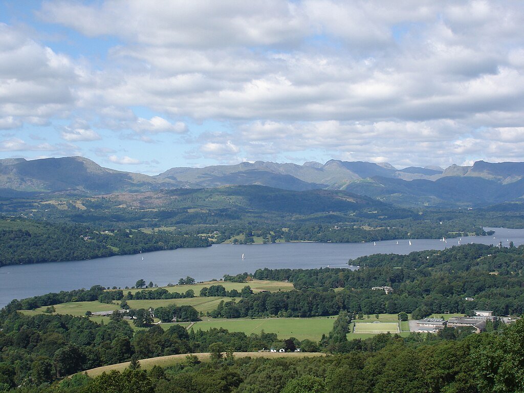 A wide scenic landscape featuring a large lake with sailboats, surrounded by green fields and forested hills, with mountains in the background under a partly cloudy sky