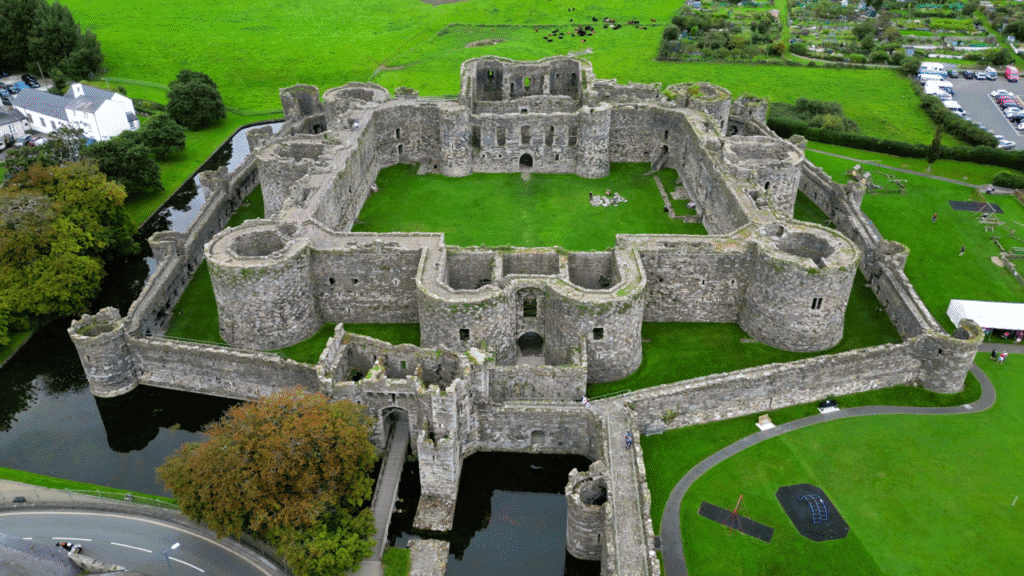 Beaumaris Castle: A Aerial view of a large, historic stone castle surrounded by a moat with green grass inside and outside the walls, situated near trees, roads, and a parking area.