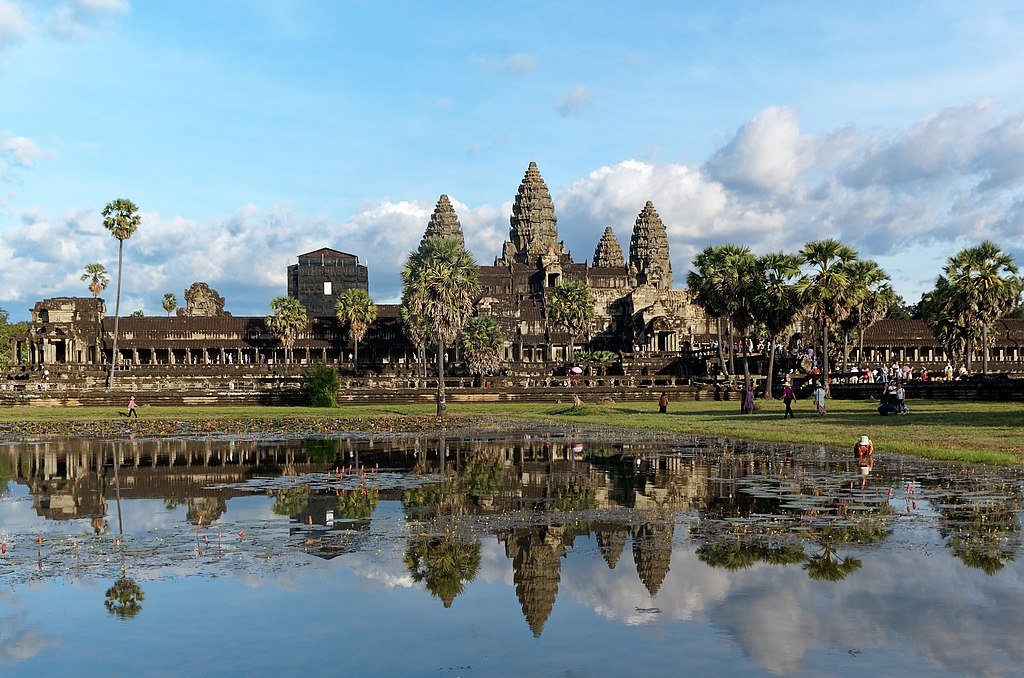 Angkor Wat temple complex reflected in a large pond with blue sky and scattered clouds, surrounded by palm trees and visitors walking nearby