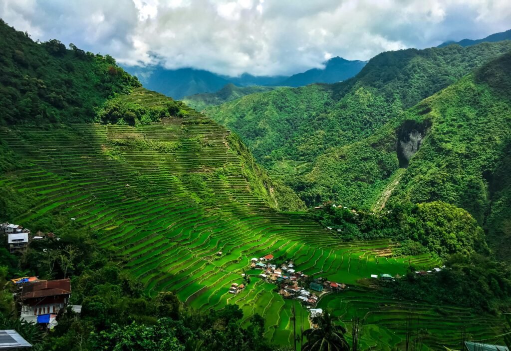 Vibrant green rice terraces on steep hills surrounding a small village, under a cloudy sky with distant mountain ranges in the background.
