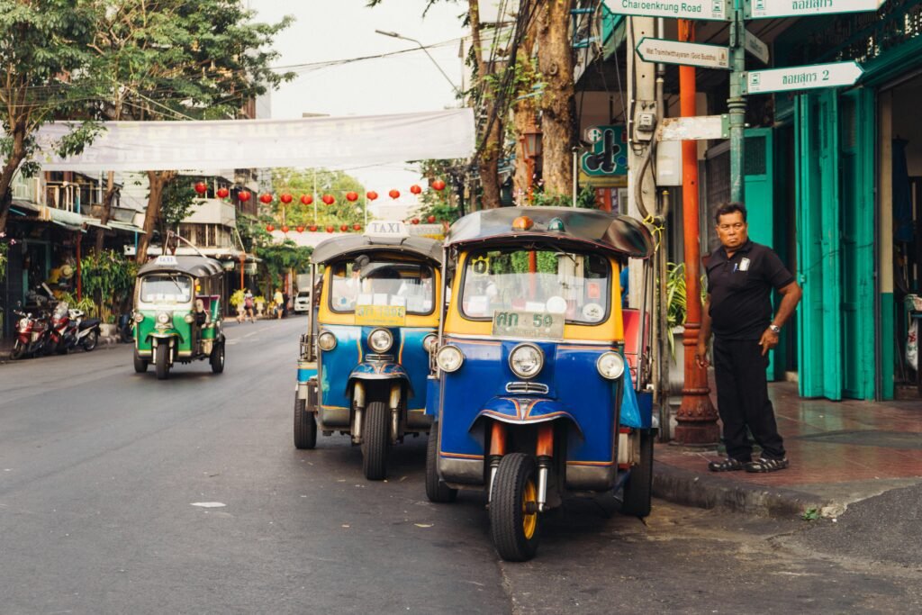 Bangkok: A Street scene with three colorful tuk-tuk taxis parked along the roadside and one tuk-tuk driving on the road, a man in black standing on the sidewalk near turquoise storefronts and various street signs overhead.