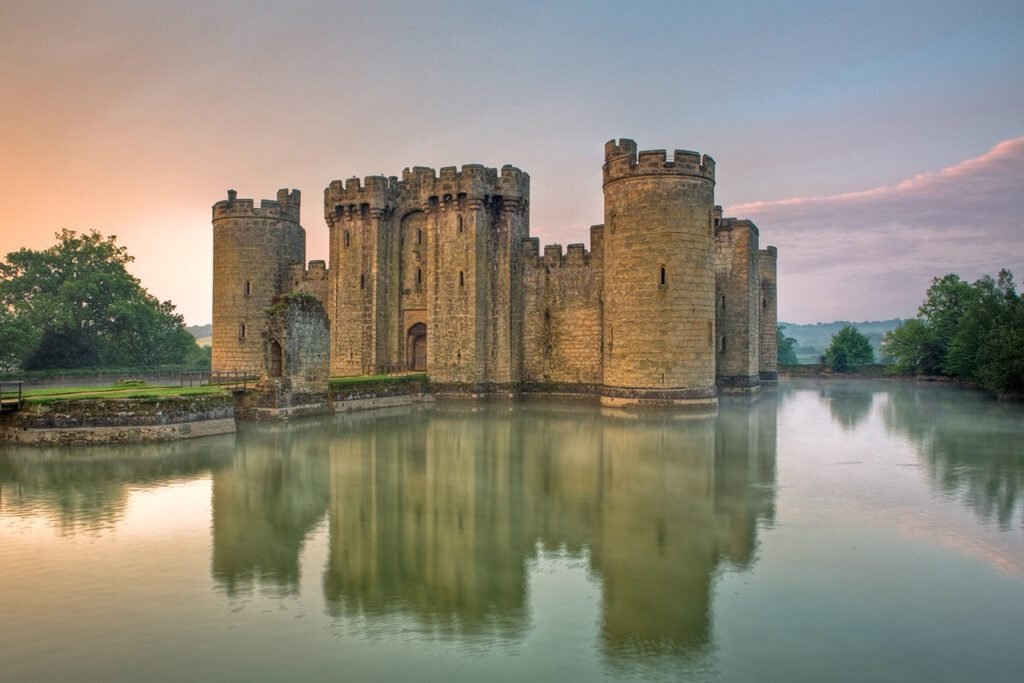 Medieval stone castle with round towers surrounded by a moat reflecting the structure, with trees and a soft pastel sky at sunrise or sunset.