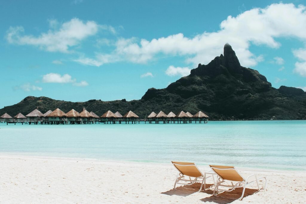 Bora Bora travel guide: Two lounge chairs and a small table on a sandy beach with calm turquoise water in front, overwater bungalows in the middle distance, and a mountain with a prominent peak under a partly cloudy blue sky.