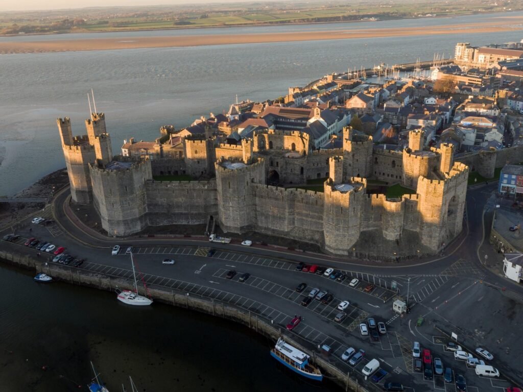 Caernarfon Castle: A Aerial view of a large medieval stone castle with multiple towers, situated next to a parking lot and waterfront, with a town and river in the background under soft evening light.