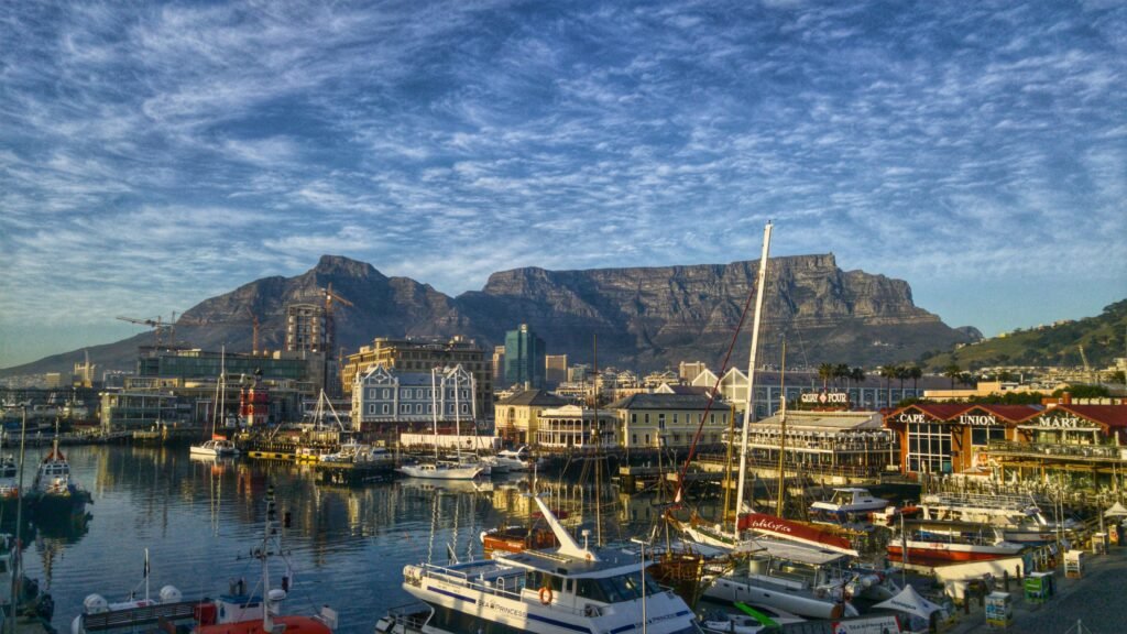 Cape Town: A View of a marina with several docked yachts and boats in front of a cityscape, with large mountains in the background under a partly cloudy sky.