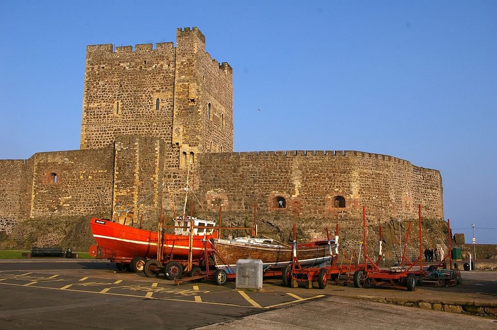 Carrickfergus Castle: Stone fortification with a large square tower and crenellated walls behind several small boats on trailers in a paved parking area under a clear blue sky