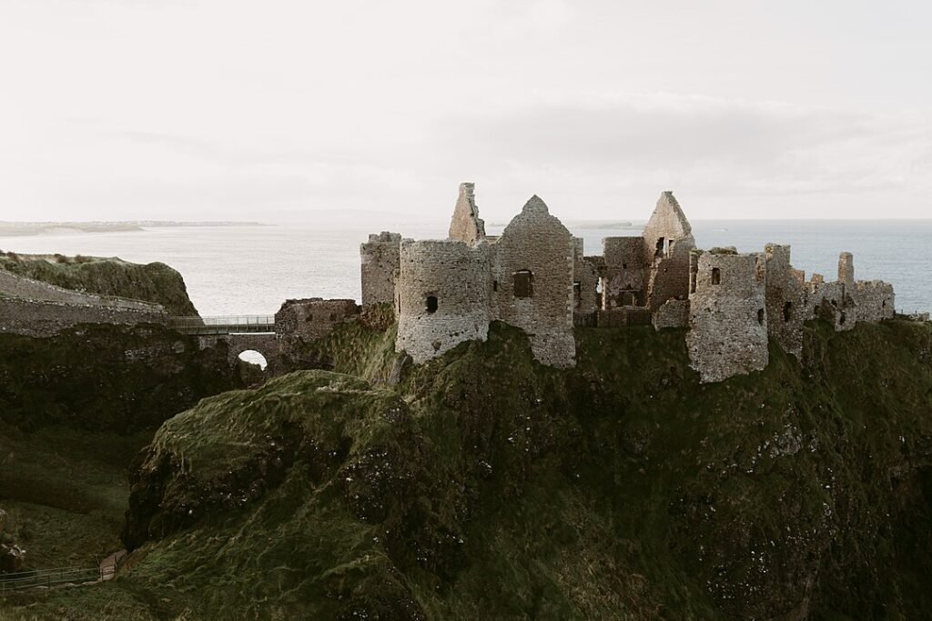 Dunluce Castle: The Ruins of a stone castle perched on a grassy cliff overlooking the sea under a cloudy sky