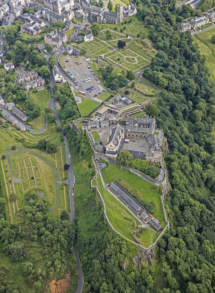 Stirling Castle: A aerial view of a large historic castle complex surrounded by green trees and lawns, with adjacent roads, parking areas, and nearby buildings in a semi-rural setting.