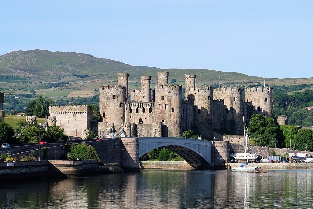 Conwy Castle: A Medieval stone castle with multiple round towers behind a stone bridge over calm water, with green hills in the background under a clear blue sky