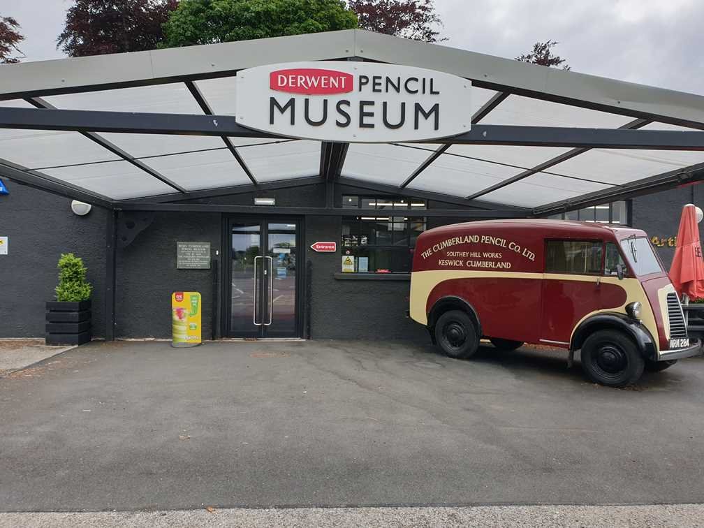 Entrance of the Derwent Pencil Museum with a covered walkway, glass double doors, and a vintage maroon and cream Cumberland Pencil Co. Ltd. delivery van parked to the right.