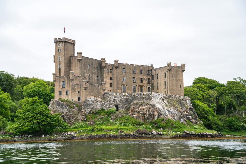 Dunvegan Castle: Historic stone castle with battlements and towers situated on a rocky, grass-covered hill surrounded by lush green trees and water in the foreground under an overcast sky.