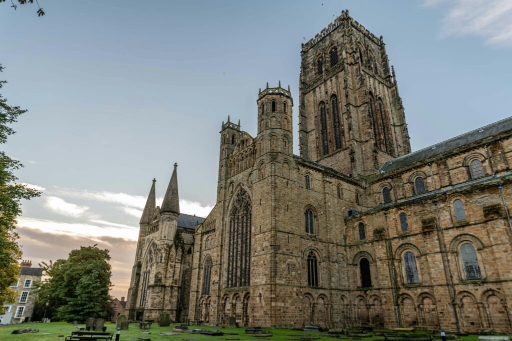 Durham Cathedral: A Large historic stone cathedral with tall towers and arched windows, set against a clear sky with nearby trees and a graveyard in the foreground.