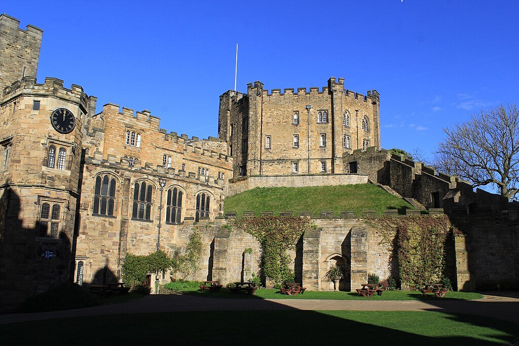 Durham Castle: A Stone castle with battlements, large arched windows, a clock tower on the left, and picnic benches on grass in the courtyard under a clear blue sky