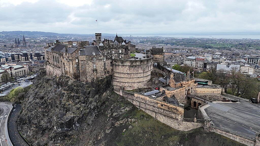 Edinburgh Castle: Scotland’s Iconic Fortress: A Aerial view of Edinburgh Castle situated on a rocky hill with historic stone walls and buildings, overlooking the cityscape under a cloudy sky.