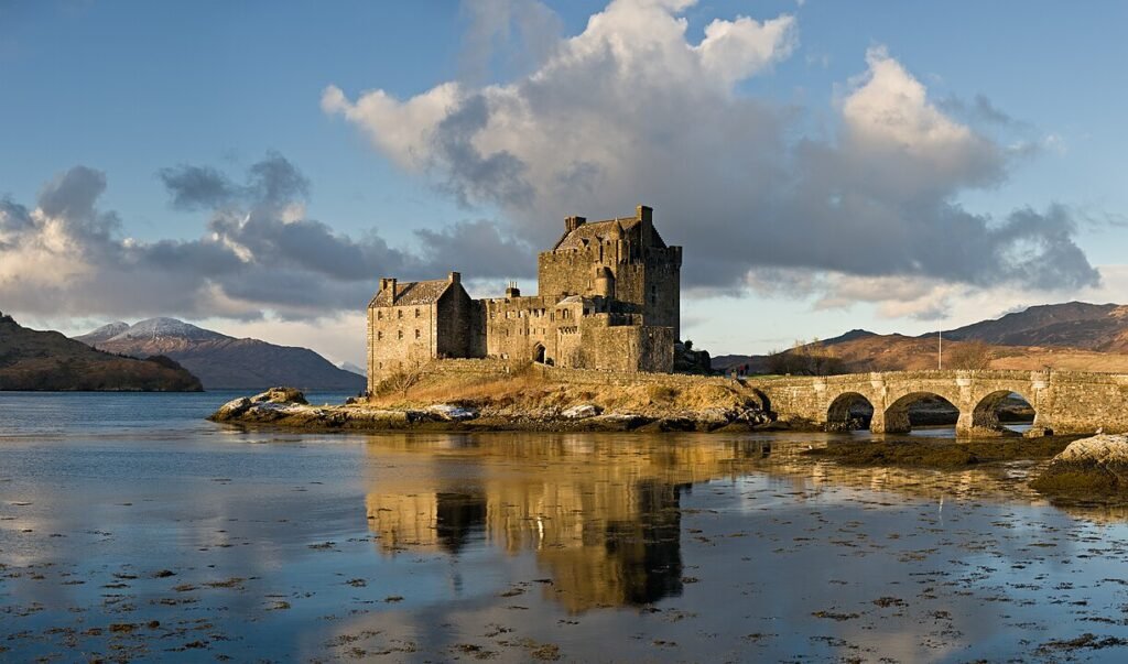 Eilean Donan Castle: A Historic stone castle on a small island connected to the mainland by a stone bridge, with calm water reflecting the structure and clouds in the sky above mountainous landscape.