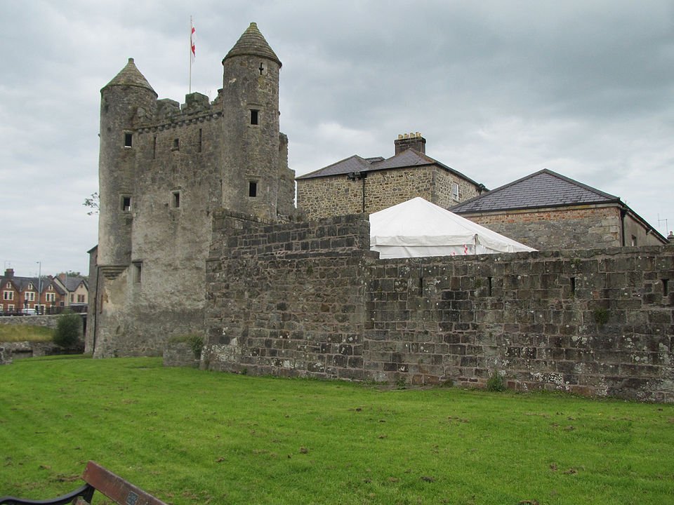 Enniskillen Castle: A Stone castle with two round towers and battlements, surrounded by a stone wall and green grass, under a cloudy sky with a white tent visible inside the walls
