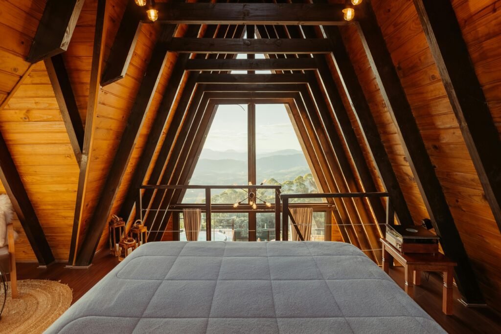Top Glamping Sites in Cumbria for a Luxury Nature Break: The Interior of a wooden A-frame cabin bedroom featuring a large window with a view of mountains, a bed with a light blue quilt, and warm lighting.