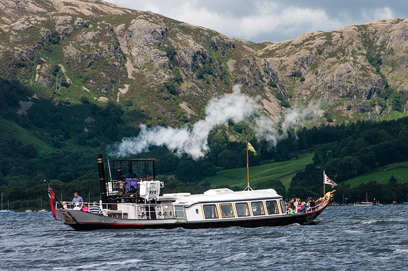 Steamboat emitting white smoke sailing on a lake with passengers on board, set against a backdrop of green hills and rocky mountains under a cloudy sky
