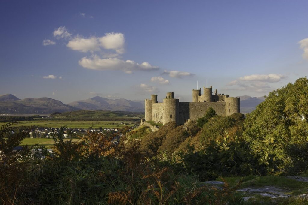 Harlech Castle: A Medieval stone castle with multiple round towers on a hill surrounded by trees, overlooking a green valley with houses and distant mountains under a clear blue sky with scattered clouds.