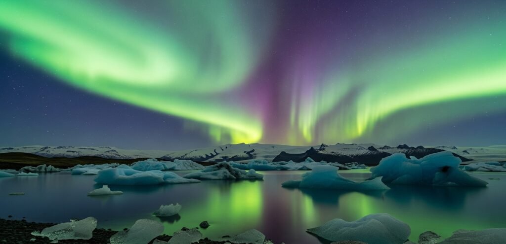Icebergs floating on a calm body of water under a night sky illuminated by green and purple northern lights with snow-covered mountains in the background