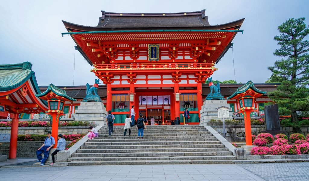 Kyoto: A Large traditional Japanese shrine gate painted in bright red with stone steps leading up to it, two guardian fox statues on pedestals, ornate lanterns, and people ascending or sitting on the steps, surrounded by flowering bushes and trees under a clear sky