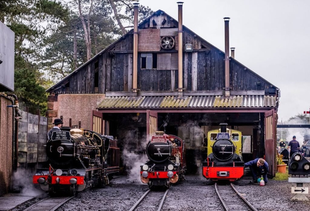 Three small steam locomotives on railway tracks in front of a rustic wooden engine shed, with two railway workers attending to the trains and steam rising around them.