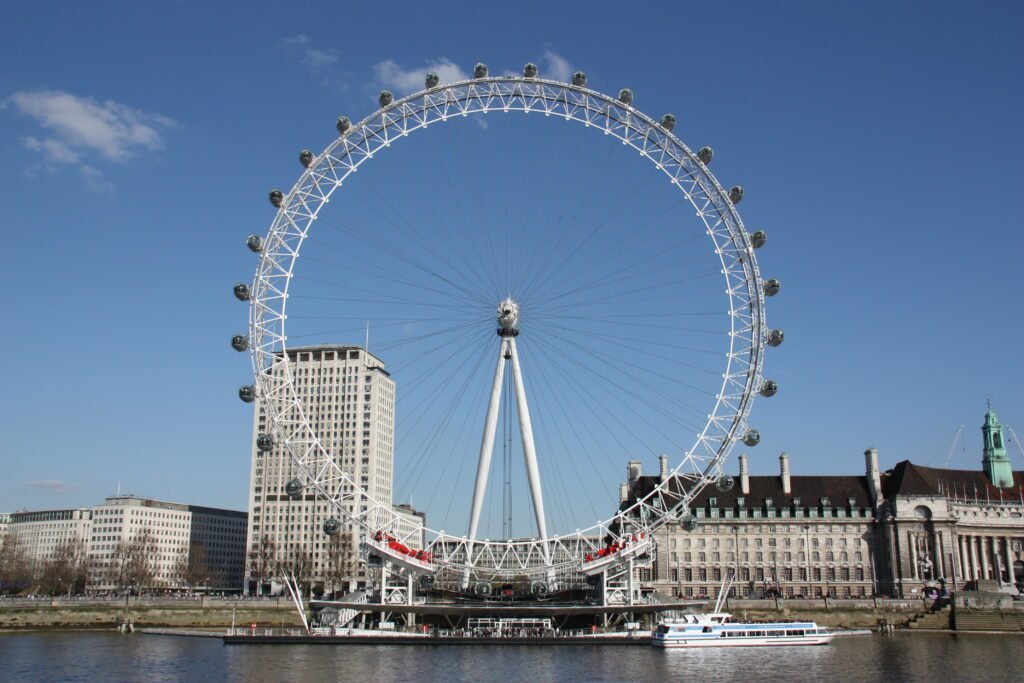 UK solo travel beginners: The London Eye Ferris wheel on the South Bank of the River Thames in London, with surrounding buildings and a boat on the river under a clear blue sky