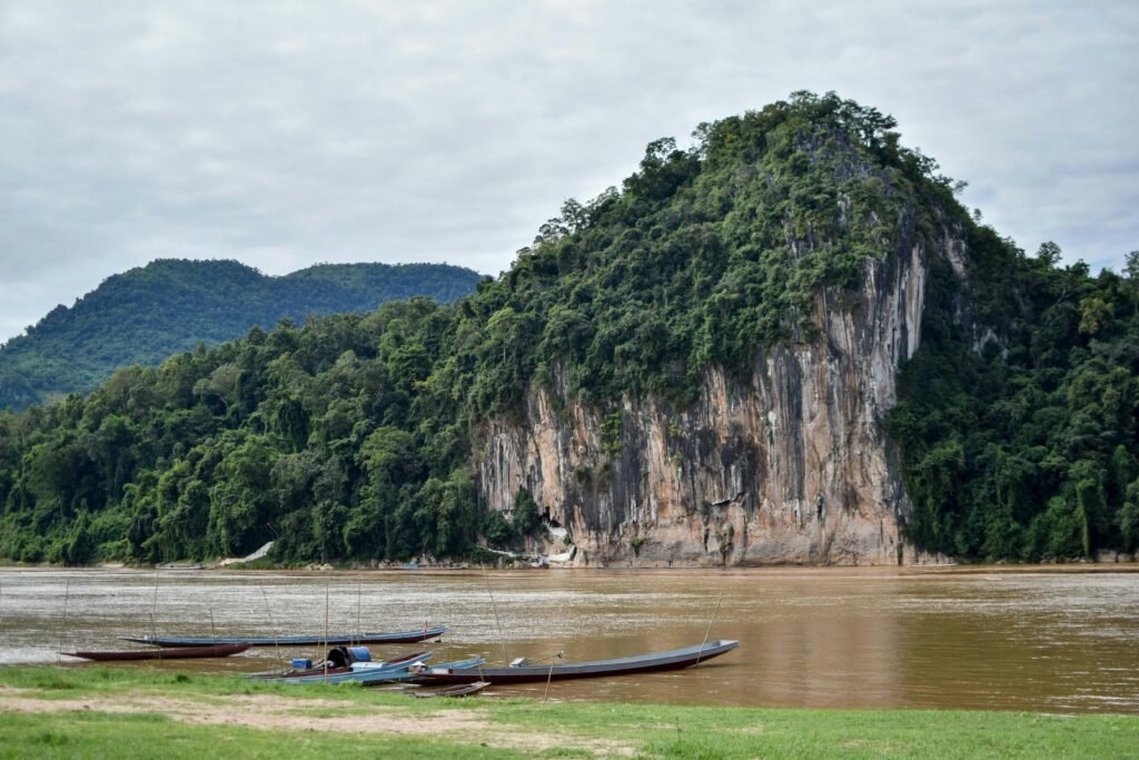 Luang Prabang: A Brown river flowing beside a forest-covered rocky hill with small boats docked on the grassy riverbank under a cloudy sky