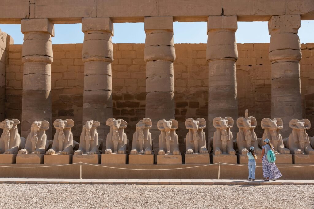 Luxor: A Row of ancient Egyptian ram-headed stone statues lined up in front of large stone columns with a woman and a child observing them outdoors under a clear sky.