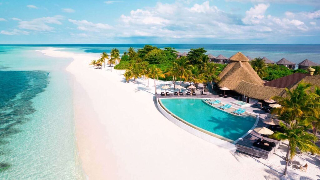 Best Time to Visit the Maldives by Budget and Weather: A tropical beach resort with white sand, palm trees, and turquoise ocean water, featuring a large swimming pool surrounded by sun loungers and thatched-roof buildings under a partly cloudy sky.
