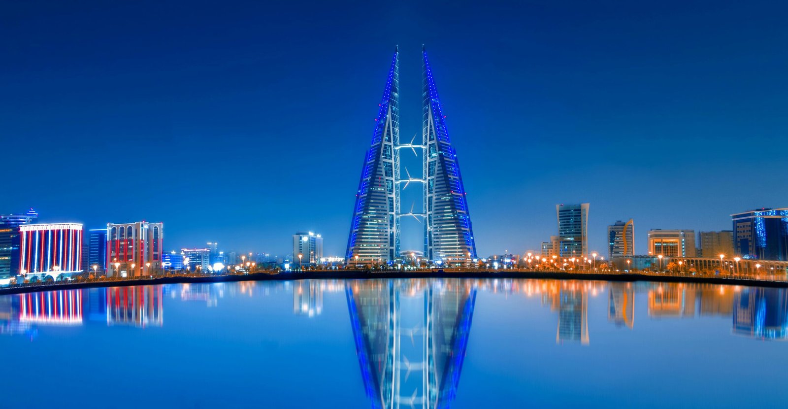 Night view of a modern city skyline with two tall, triangular skyscrapers lit with blue lights and wind turbines in between, reflecting clearly in a calm body of water in the foreground, under a deep blue sky.