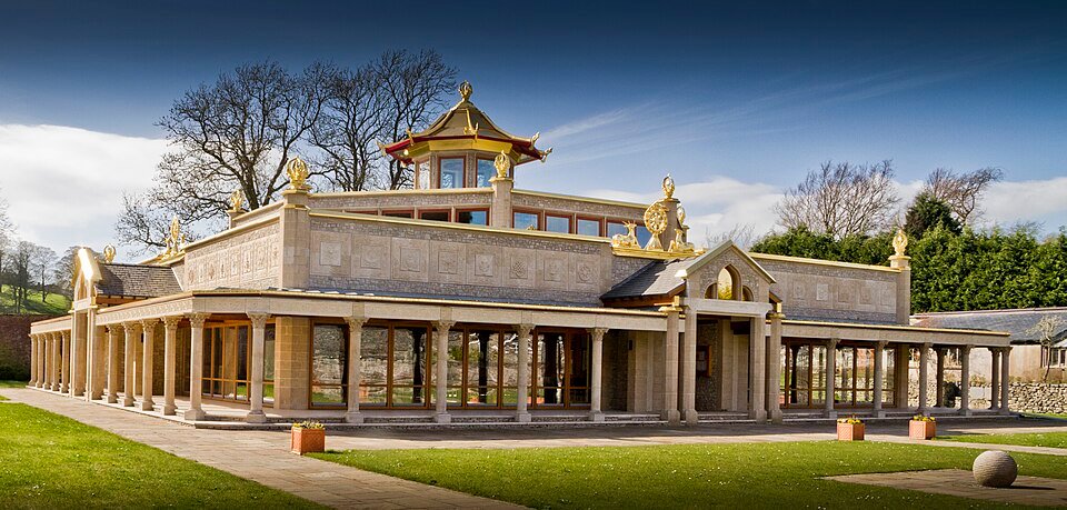 Unusual things to do in Cumbria: A large, ornate building with a flat roof surrounded by stone columns and a decorative golden pagoda-style structure on top, set in a grassy area with a paved walkway and trees in the background under a blue sky