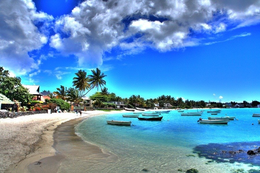 Mauritius vs Seychelles: Which Island is Right for You?: A tropical beach with clear blue water and several small boats anchored near the shore, lined with palm trees, houses, and a partly cloudy blue sky overhead.