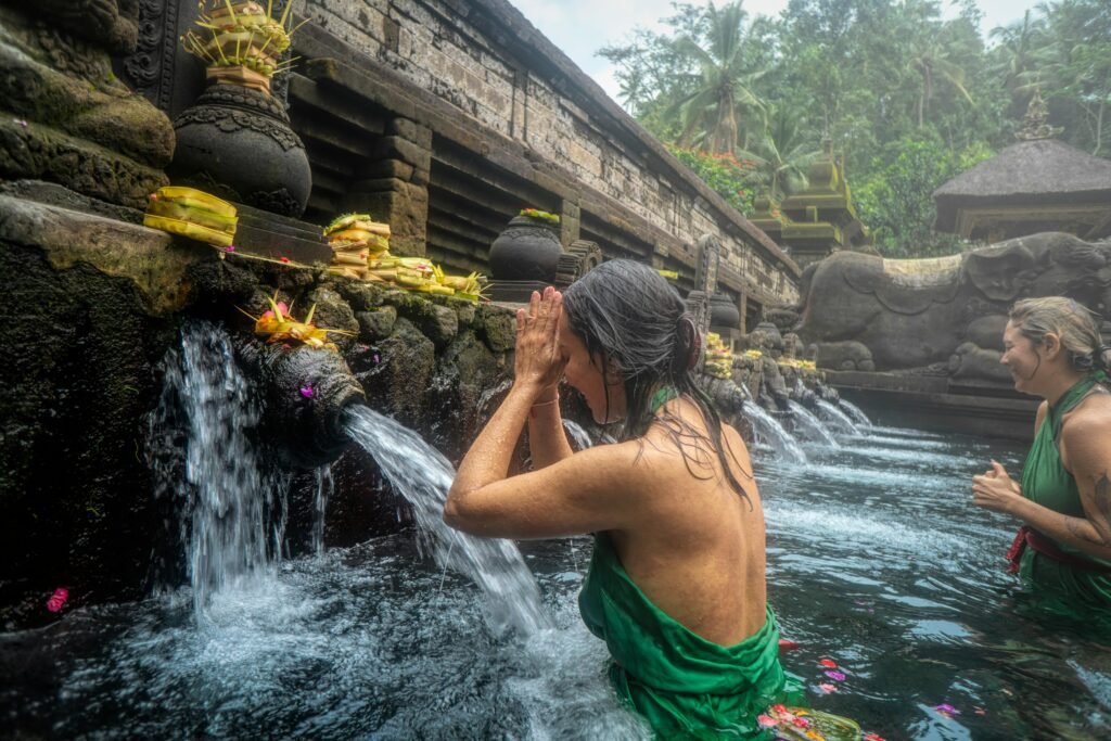 Bali Travel Guide: Two women wearing green sarongs participating in a cleansing ritual under water spouts at a traditional temple fountain surrounded by stone carvings and greenery.