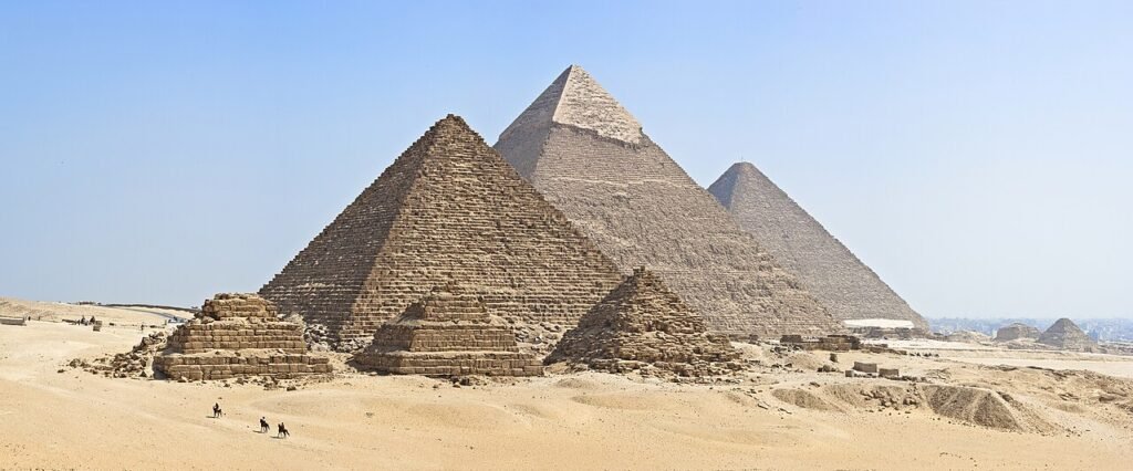 View of the pyramids of Giza in Egypt with smaller pyramids in front and three larger pyramids behind, set against a clear blue sky and desert sand, with a few people on camels in the foreground