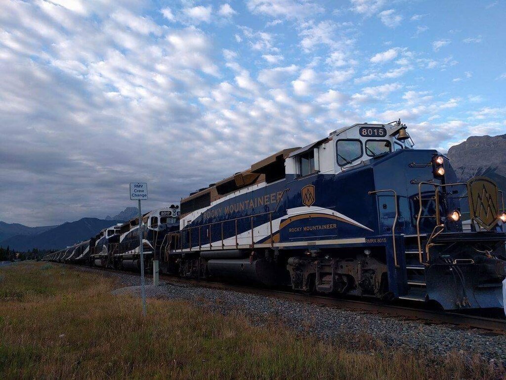 7 Epic Rail Journeys Around the World: A long Rocky Mountaineer train with blue and gold locomotives on railway tracks under a cloudy sky with mountains in the background and a sign that reads 'RMV Crew Change' beside the tracks.