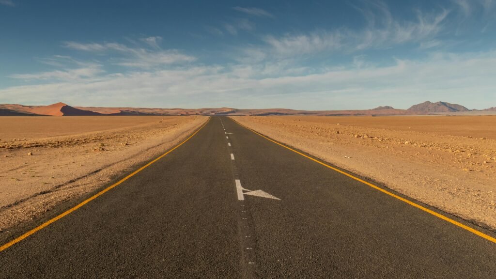 Sossusvlei Dunes: A Straight asphalt road with yellow side lines and a white arrow pointing left in the middle, stretching through a dry desert landscape with mountains in the distance under a blue sky with scattered clouds.