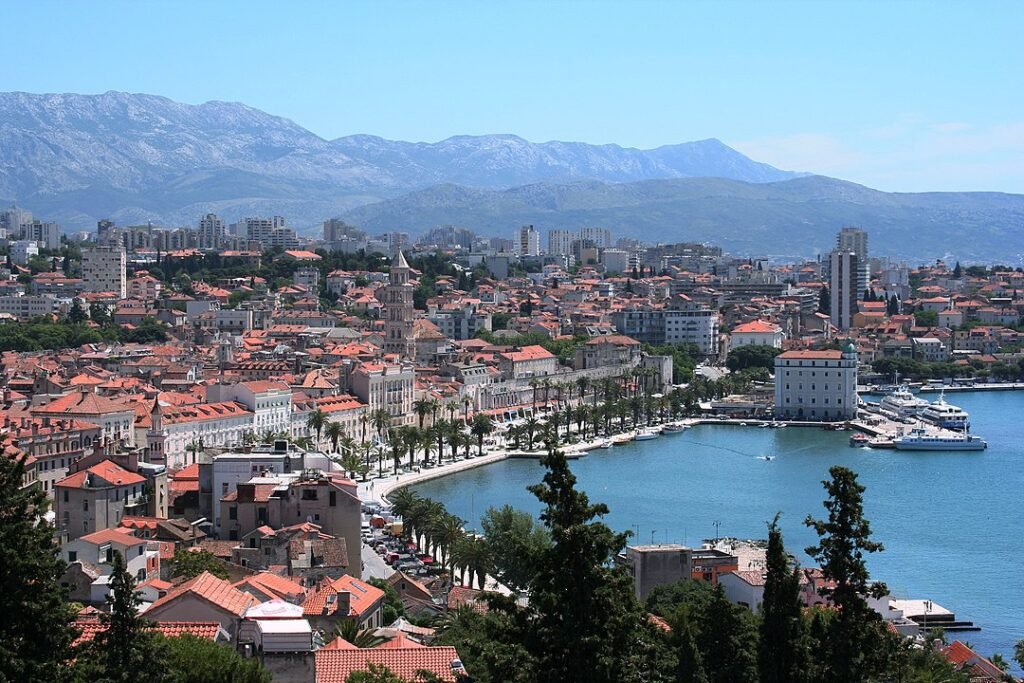 Split-Croatia: A Panoramic view of a coastal city with red-tiled roofs, a prominent bell tower, a waterfront lined with palm trees, boats docked at the harbor, and mountains in the background under a clear blue sky