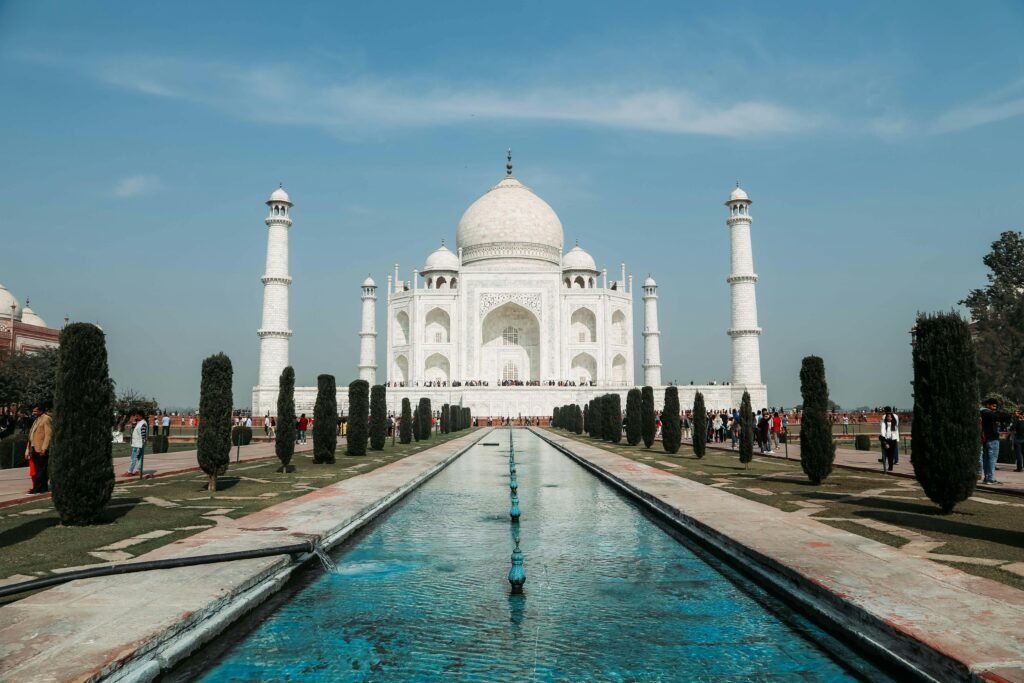 Front view of the Taj Mahal with its white marble dome and minarets, framed by symmetrical rows of tall trees and a long reflecting pool under a clear blue sky.