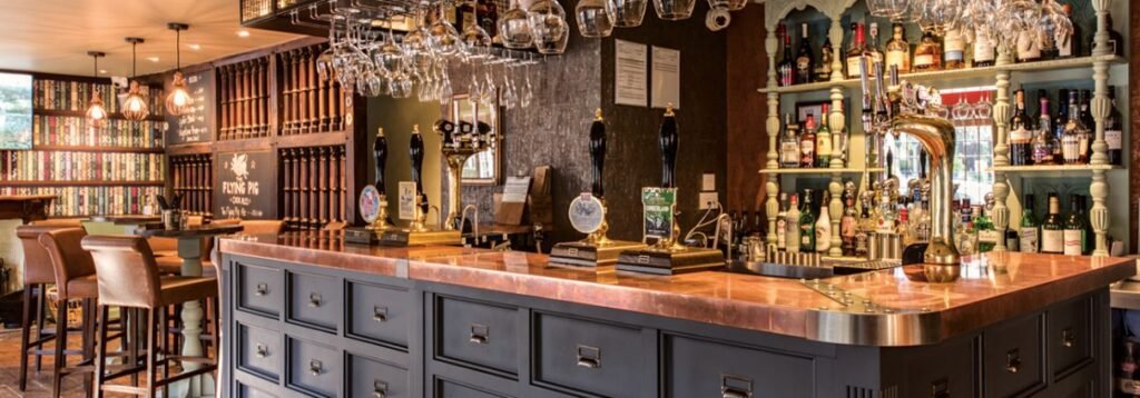 Interior of a well-stocked traditional pub bar with high stools, hanging glassware, beer taps, and shelves filled with various bottles of alcohol, featuring warm lighting and vintage decor.