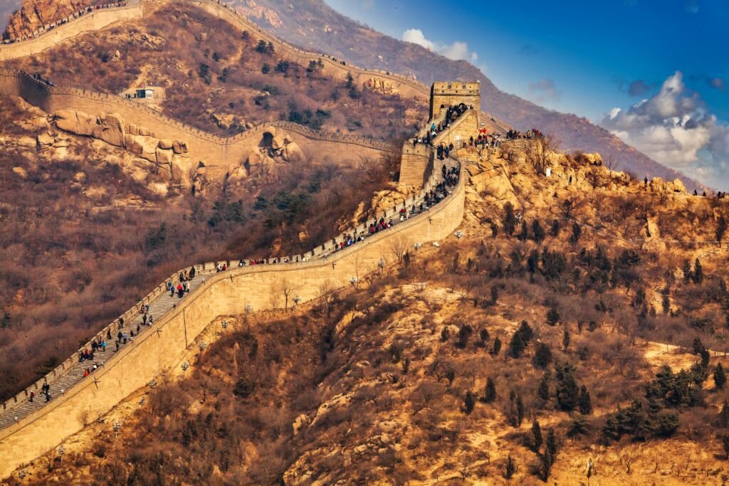 The Great Wall of China: A Section of the Great Wall of China winding over brown, rocky hills with sparse vegetation under a blue sky with some clouds and people walking along the wall.
