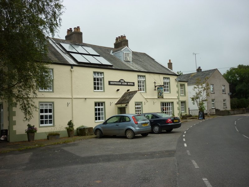 A pale yellow two-storey building with a slate roof and solar panels, identified by a sign as The Shepherds Arms Hotel, with two parked cars in front and a curved road alongside under an overcast sky.
