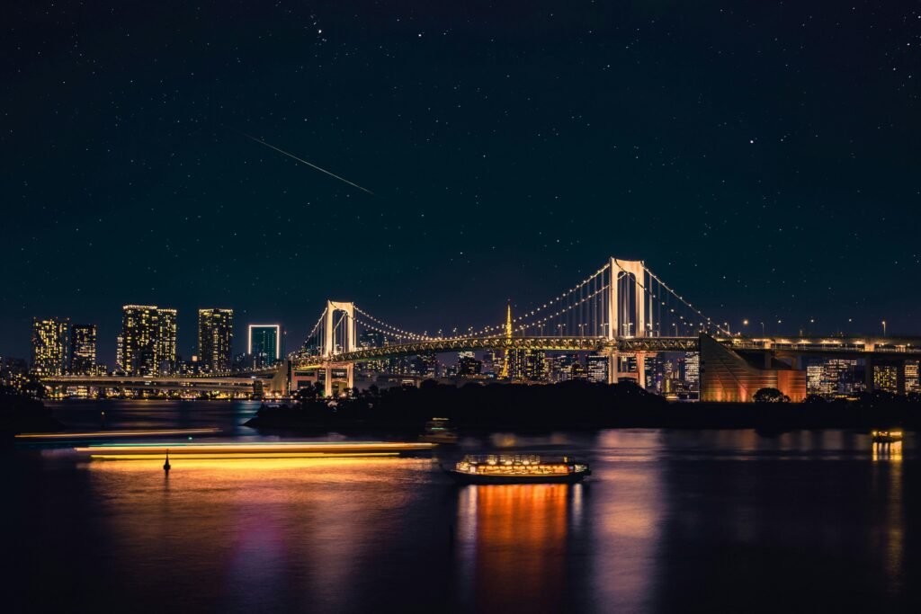 Tokyo: An Illuminated suspension bridge and city skyline at night with starry sky, reflecting on calm water with blurred lights of passing boats