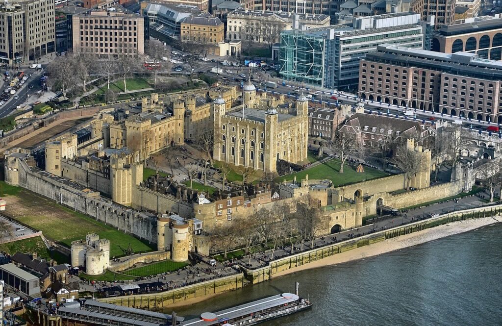 Tower of London: A Aerial view of the Tower of London surrounded by its medieval stone walls and towers, with the River Thames in the foreground and modern city buildings in the background.