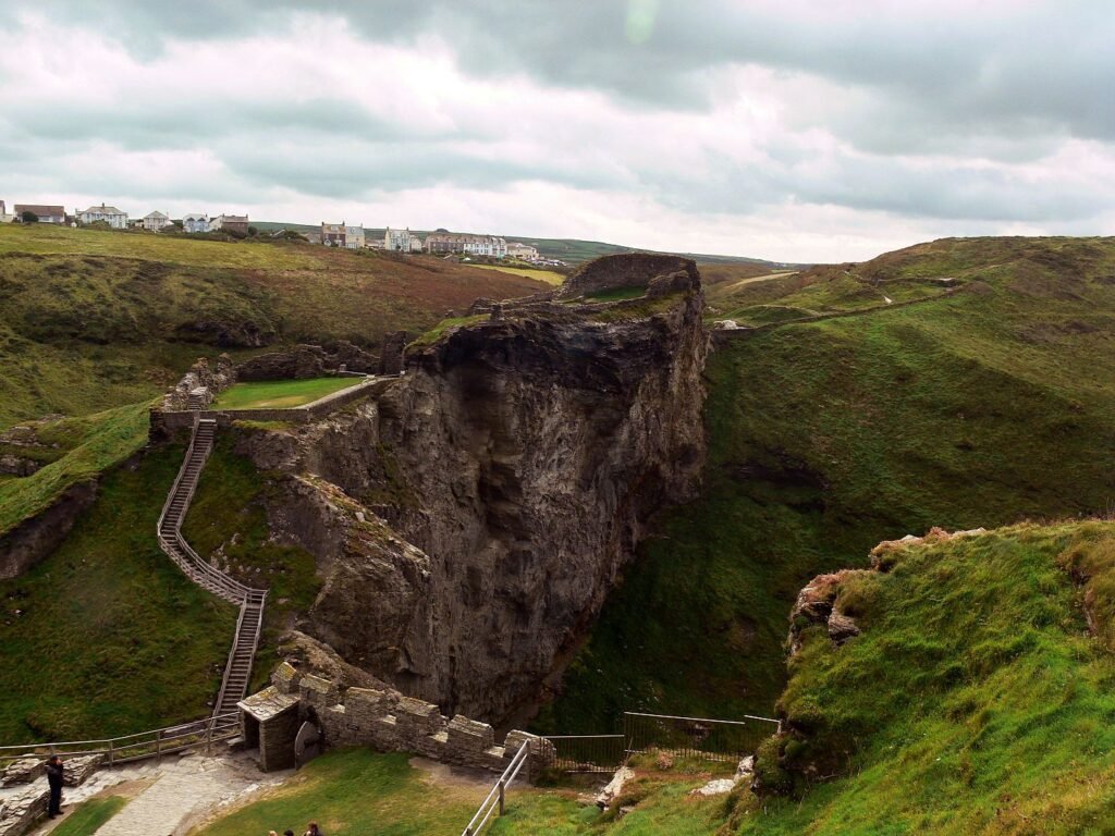 Tintagel Castle: Ruins of a stone castle situated on a rugged cliff surrounded by green hills under a cloudy sky, with a village visible in the background and a wooden staircase leading up the hill.