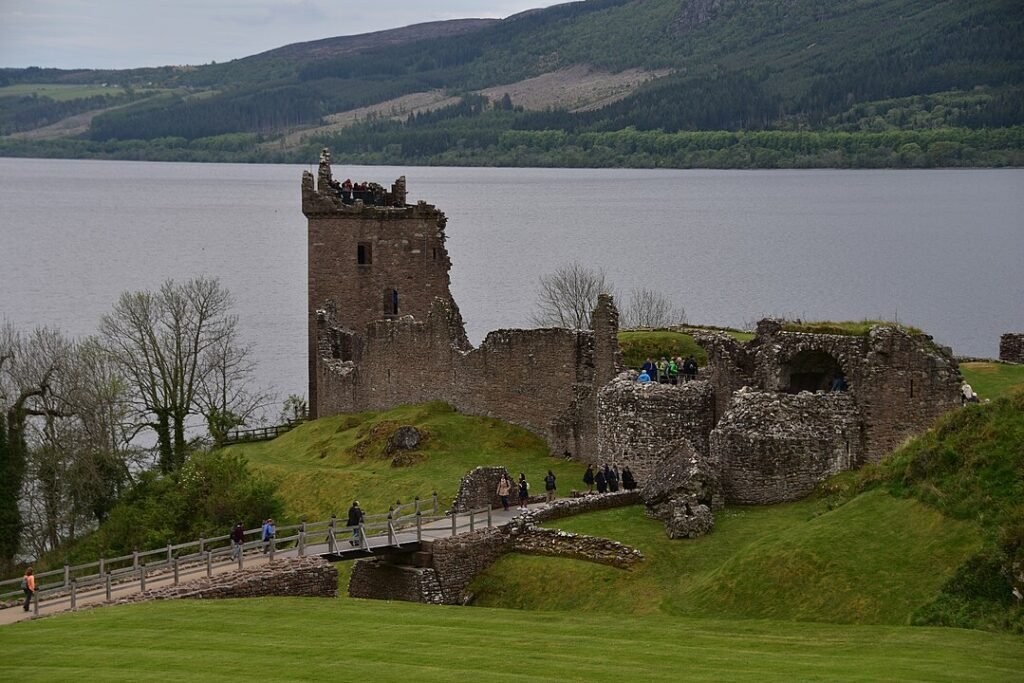 Urquhart Castle: Ruins of a stone fortress with a tall tower beside a large lake, surrounded by green hills and a wooden bridge leading to the entrance, with people walking and exploring the site.
