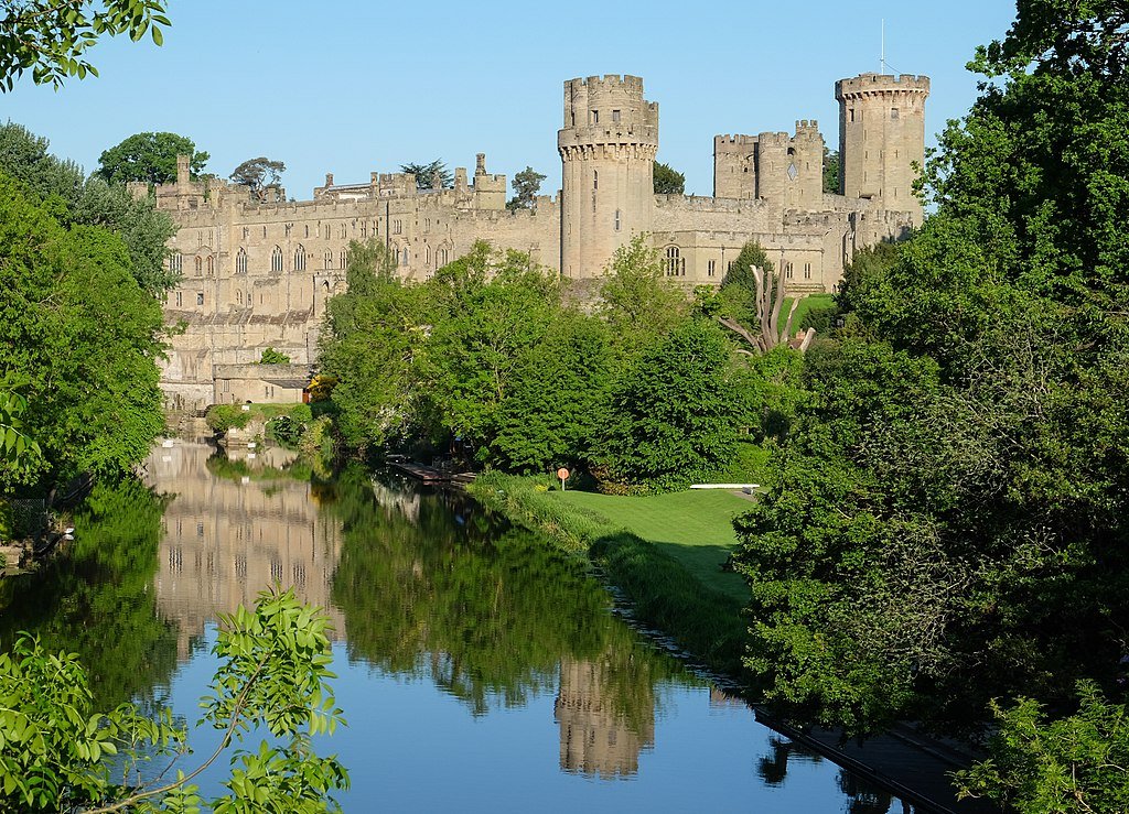Warwick Castle: A Historic stone castle with multiple towers beside a calm river reflecting the castle and surrounding green trees under a clear blue sky