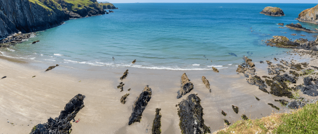 Top 10 Secret Beaches in Europe | You Can Actually Reach: A Wide sandy beach with scattered dark rocky formations extending into the shallow blue sea, steep cliffs covered in green grass on the left, and clear blue sky above.